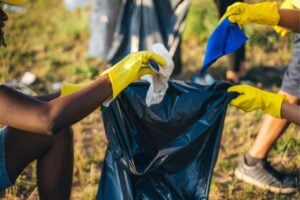 People wearing yellow gloves picking up litter and placing it into a dark blue garbage bag during an outdoor cleanup.