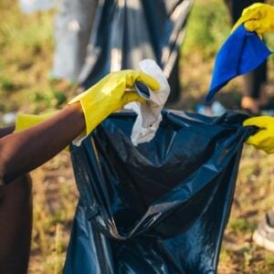 People wearing yellow gloves picking up litter and placing it into a dark blue garbage bag during an outdoor cleanup.
