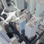 White quadcopter drone with a mounted camera flying above a dense city of tall skyscrapers, capturing aerial footage.
