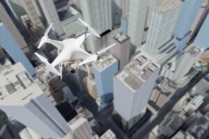 White quadcopter drone with a mounted camera flying above a dense city of tall skyscrapers, capturing aerial footage.