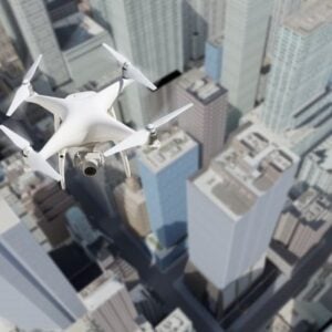 White quadcopter drone with a mounted camera flying above a dense city of tall skyscrapers, capturing aerial footage.