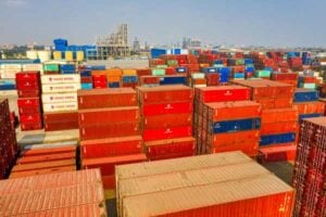 A vast shipping yard with many stacked colorful containers (red, orange, blue, white) under a clear sky; distant port buildings in the background.