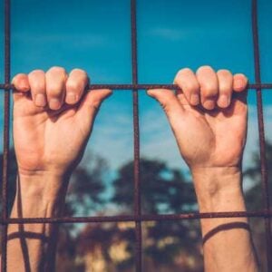Hands gripping rusted metal bars of a fence against a bright blue sky behind.