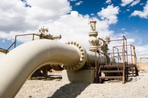 Large beige industrial pipeline with a bolted flange, valves, and metal catwalks on a gravel site under a blue sky.