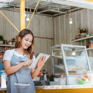 Woman in an apron uses a tablet at a cafe counter with a pastry display behind her, smiling as she works.