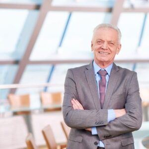 Senior businessman in a gray suit with arms crossed in a bright, contemporary office atrium.