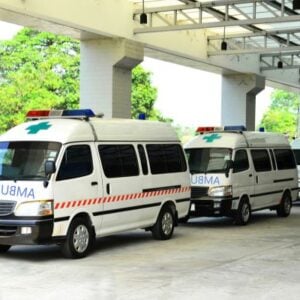 Three white ambulances with red stripes and blue emergency lights parked under a covered driveway, with green trees in the background.