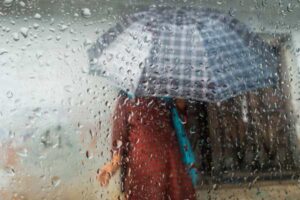 Two people share a plaid umbrella in heavy rain, seen through a raindrop-covered window.