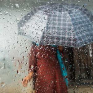 Two people share a plaid umbrella in heavy rain, seen through a raindrop-covered window.