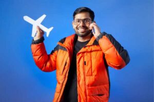 Man in an orange puffer jacket smiles while talking on a cellphone and holding a white paper airplane against a blue background.